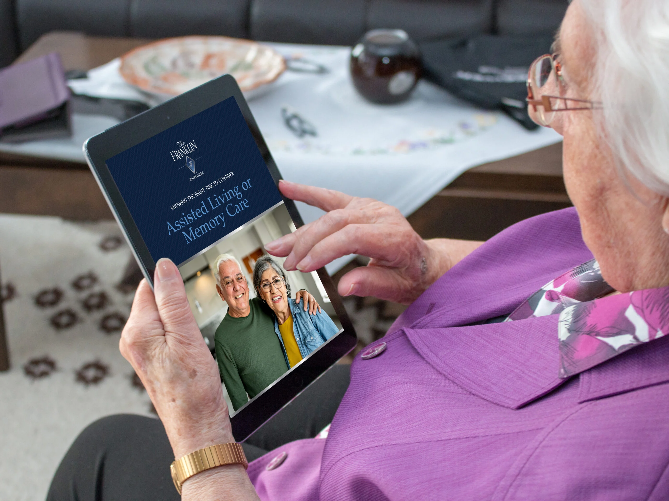 A senior resident views a tablet displaying information about assisted living and memory care, in an indoor community setting.